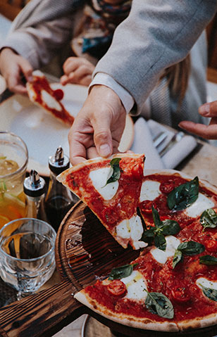 Hand picking up a slice pizza with mozzeralla and spinach. Table is set with glasses, condiments and a blurred vision of someone holding a slice of pizza over a plate.