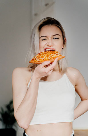 A blonde female taking a bite from a slice of pizza and winking at the camera.