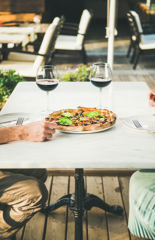 Table set with two wine glasses halfed filled with red wine, a pizza on a plate in the middle and a set of male hands on the table. Blurred background of other empty chairs and tables.