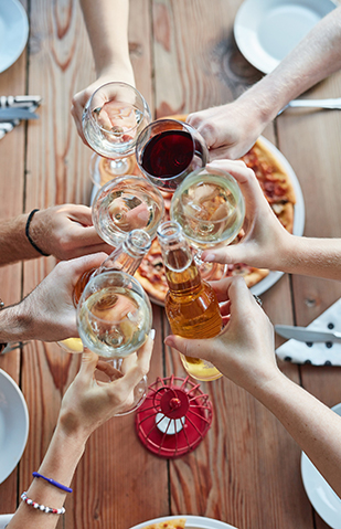 shot from above, display hands cheering with beer bottels and glasses of white and red wine. Table set with white plates, cutlery and napkins with a blurred image of a pizza in the middle.